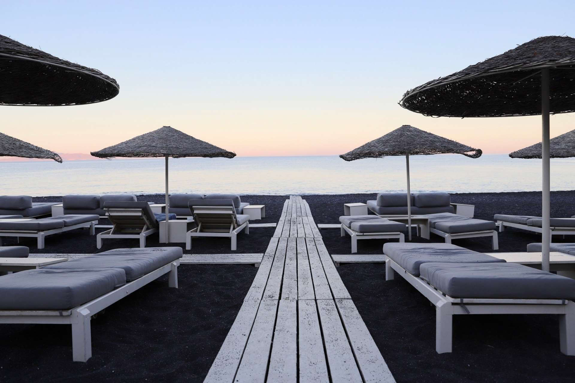 Solsenger og parasoller p&aring; svart sandstrand i Kamari p&aring; Santorini med rolig hav