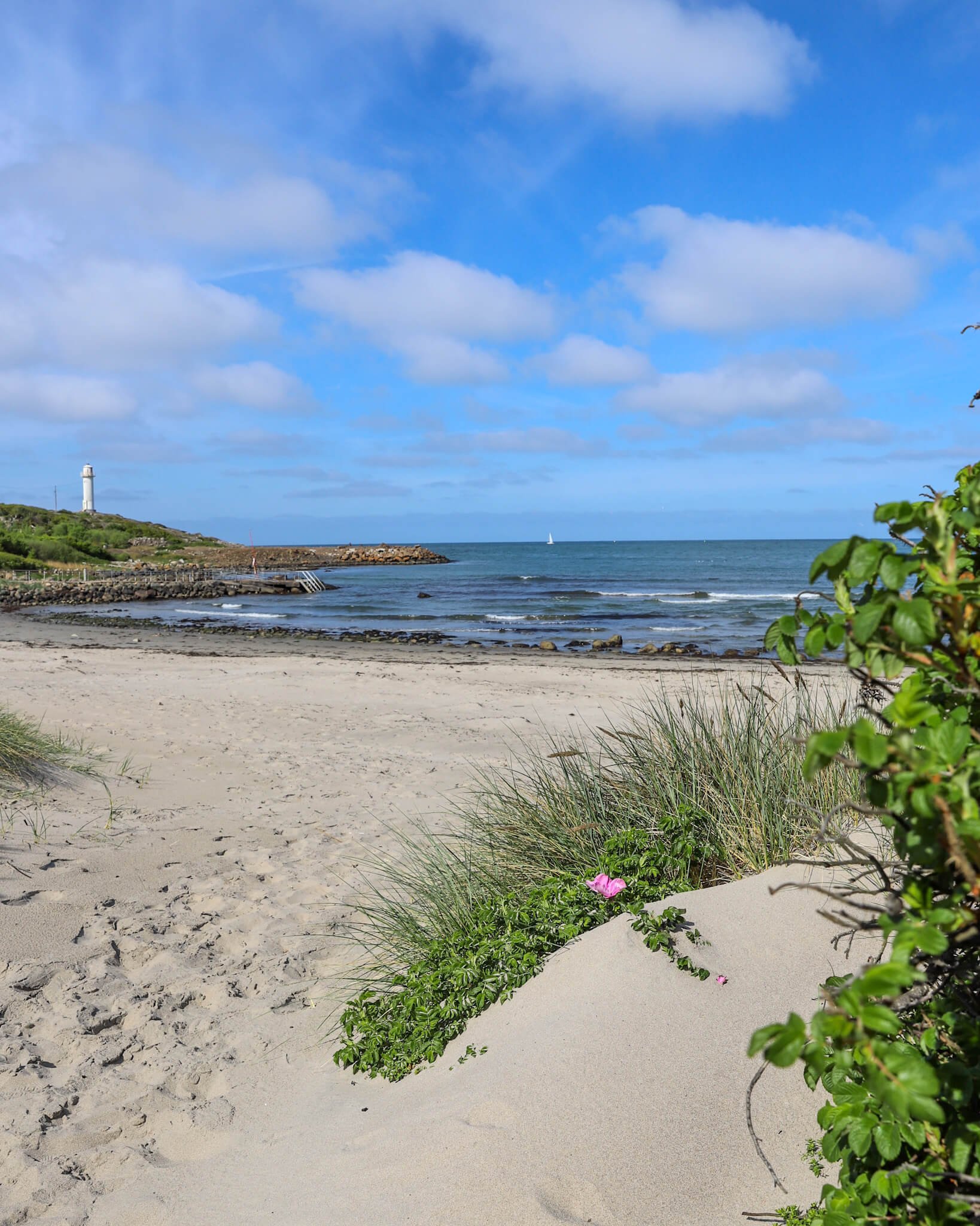 Lys og fin sandstrand, havet med bølger som slår mot land og Subbe fyr i Lilla Apelviken. 