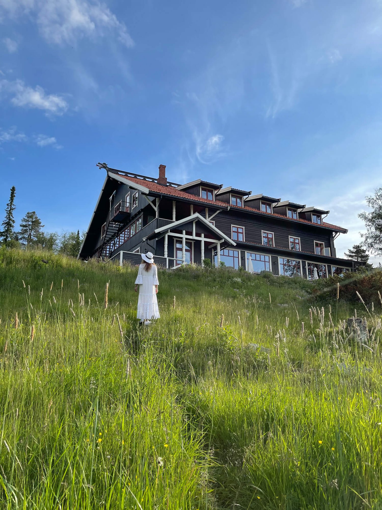 Dame med hvit kjole og hvit hatt går gjennom gresset mot Tuddal Hoyfjellshotell. Det er sommer og blå himmel. 