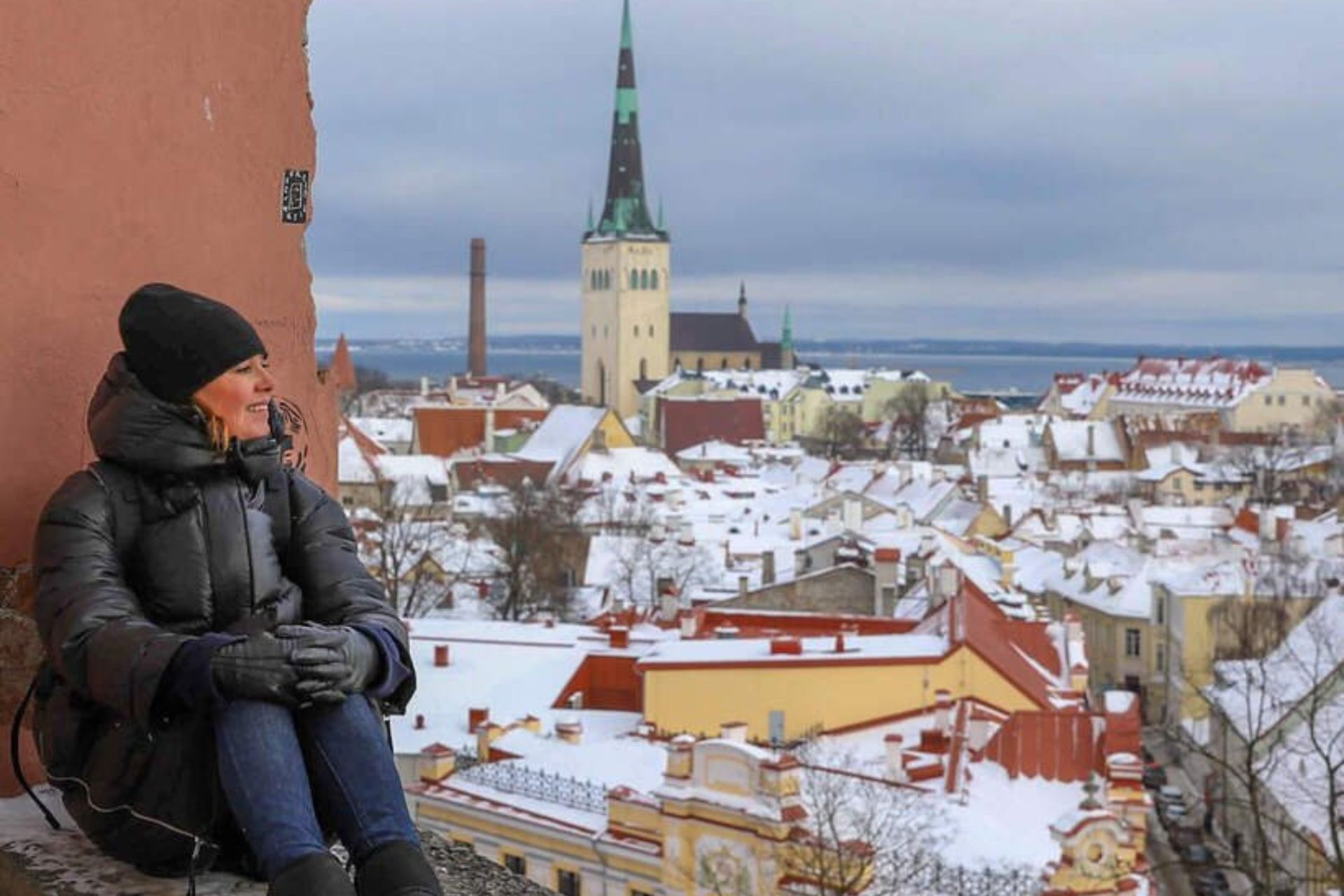 Dame i vinterklær sitter ved bymuren med utsikt over Tallinns snødekte gamleby og St. Olavs kirke.