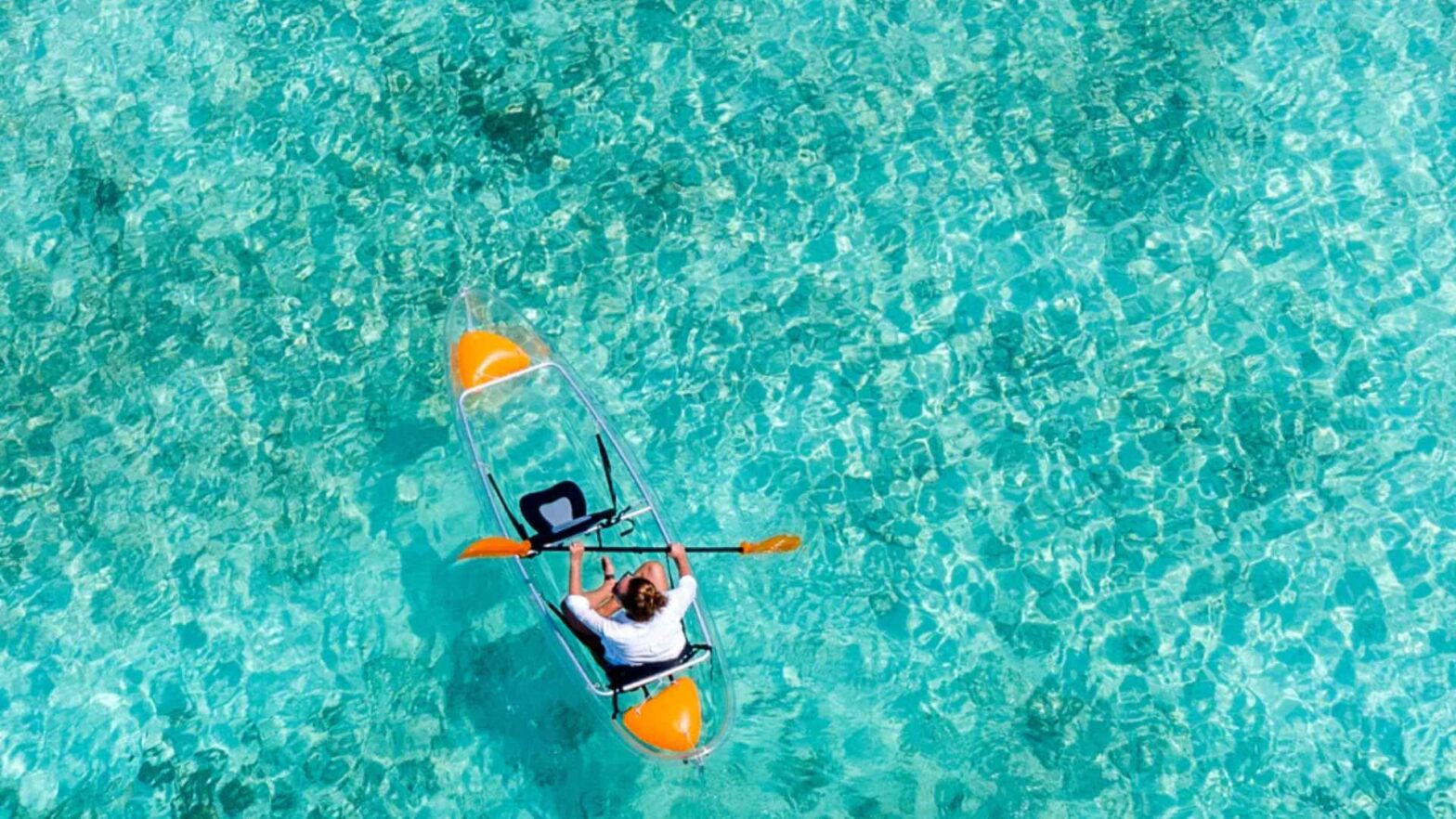Dronebilde av dame som padler i glasskajakkk på turkist hav på Maldivene.