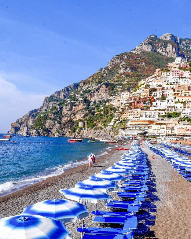 Blå solsenger, blå og hvite parasoller, blått hav og Positano by sett fra stranden.