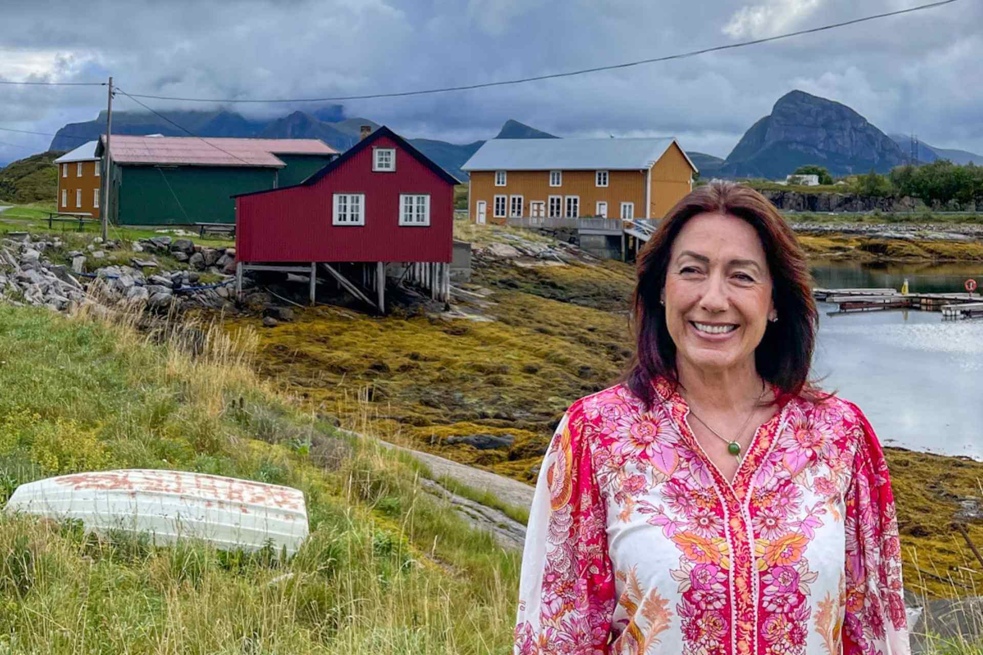 Kystlandskap på Helgelandskysten med kvinne foran rorbuer, fjell og rolig sjø.