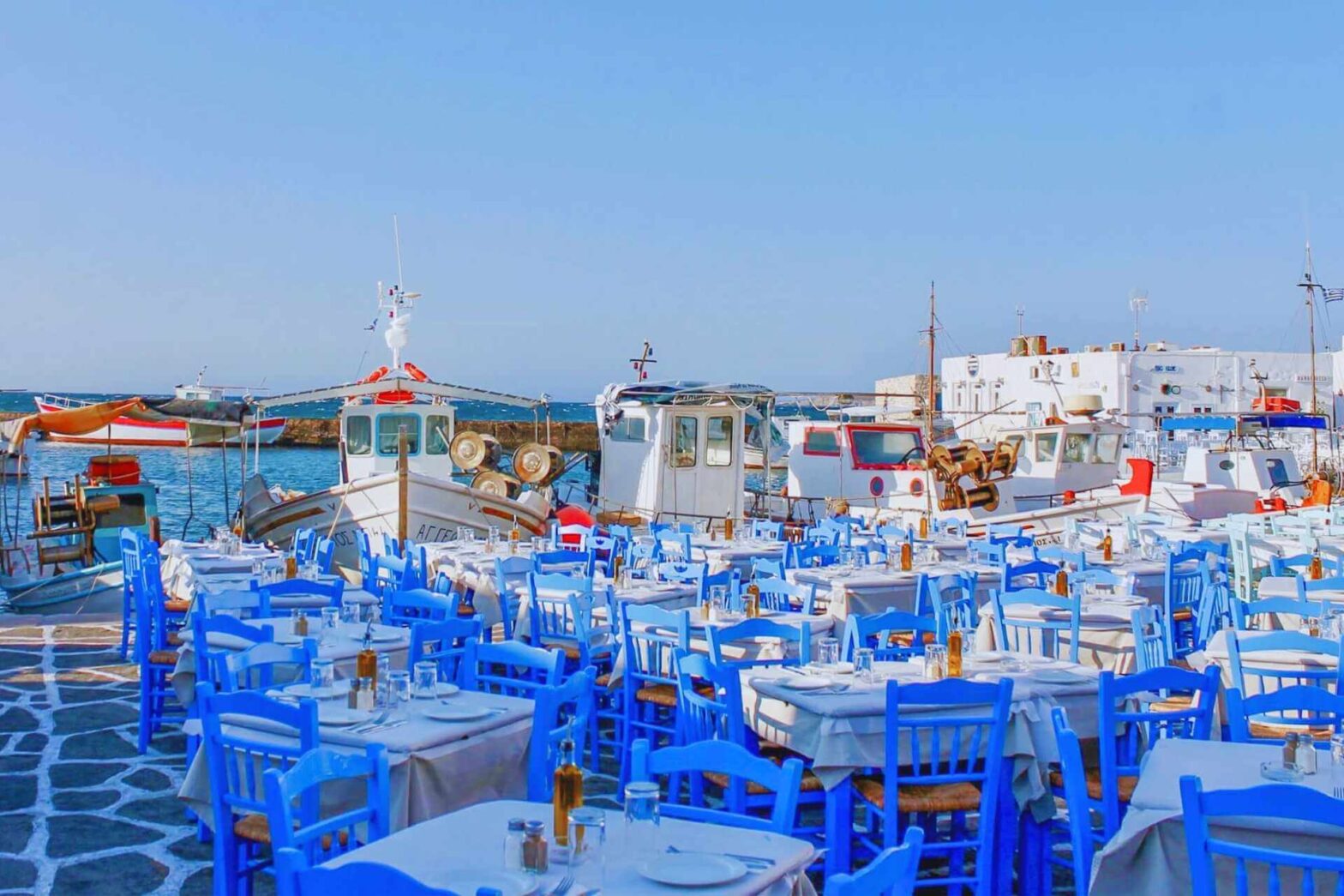 Uteservering med blå stoler ved havnen i Naoussa på Paros, omgitt av fiskebåter og hvitkalkede hus.
