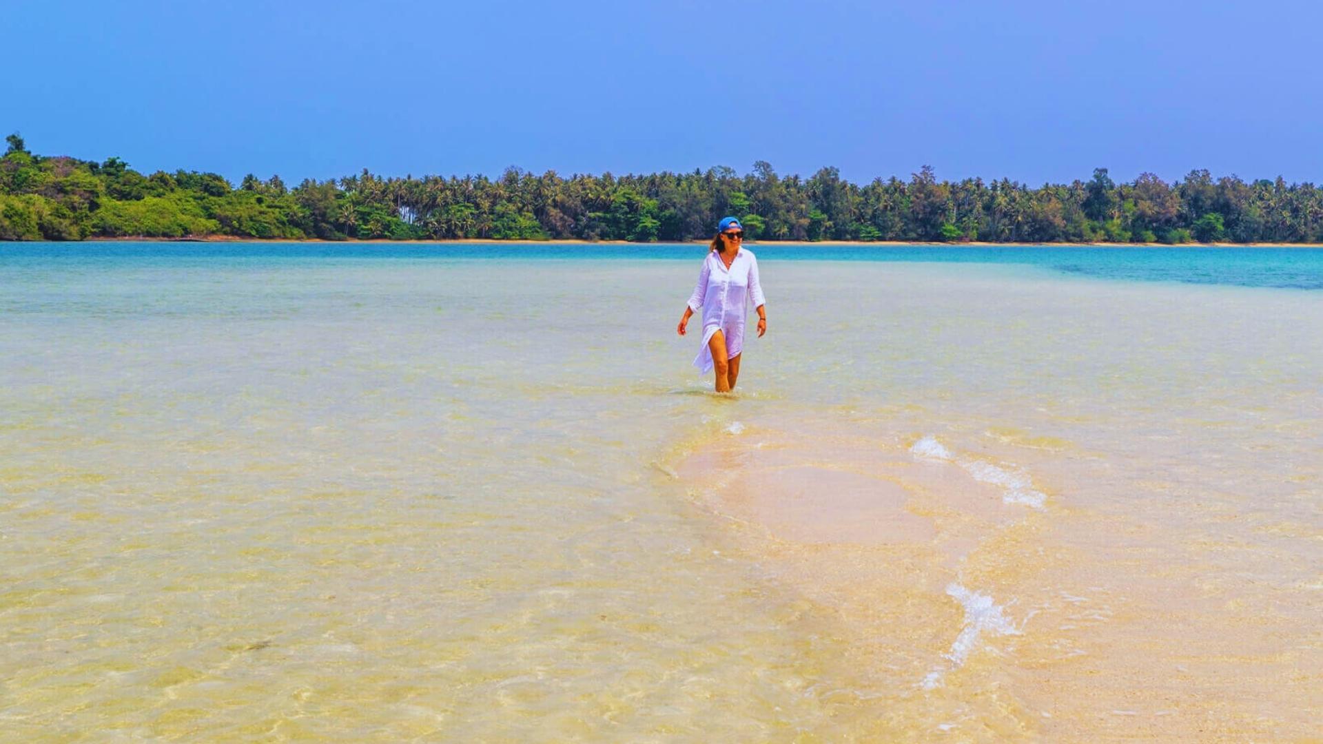 Kvinne som går langs vannkanten på en stille strand nær Koh Mak i Thailand, omgitt av klart turkist hav og gyllen sand. Foto: Merete Moen.