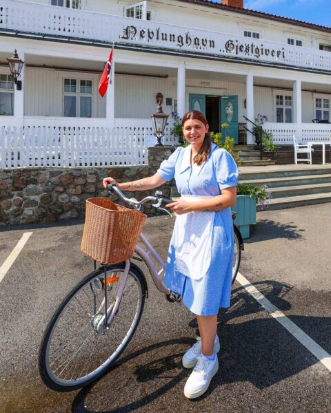 Smilende kvinnelig ansatt i gammeldags lyseblå uniform og damesykkel med kurv utenfor Nevlunghavn Gjestgiveri.