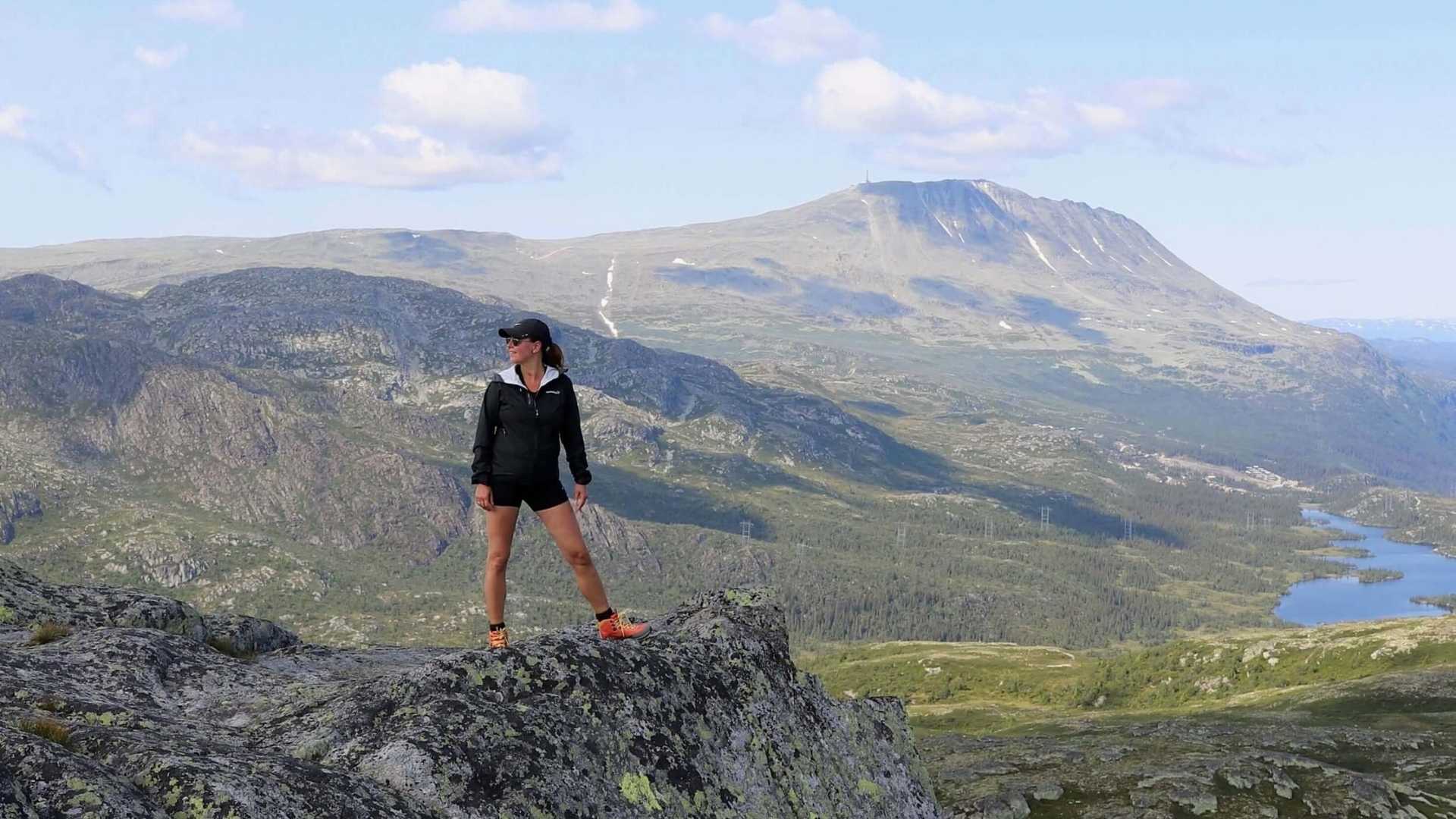 Gaustatoppen sett fra Ørnenipa. Sprek dame i sorte turklær står på Ørnenipa med Gaustatoppen i bakgrunnen. 