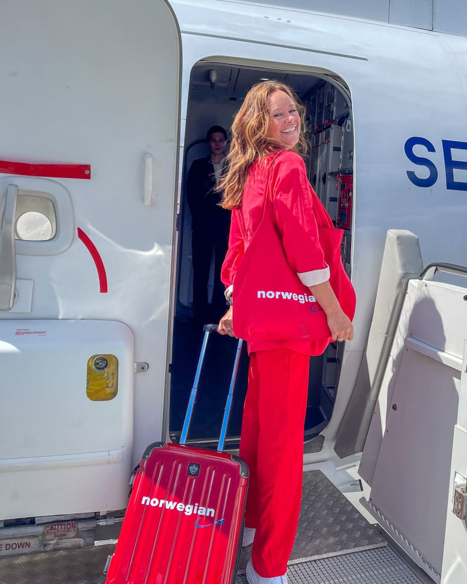 Smilende dame i rød dress med rød koffert fra Norwegian på vei inn på et Norwegian-fly.