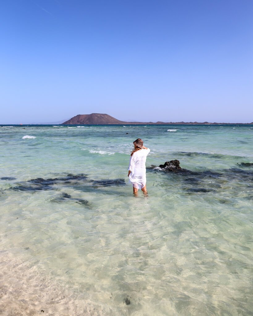 Her står jeg i hvit lang skjorte, med sjøen opp til anklene på en strand på Fuerteventura og ser mot øya Los Lobos.