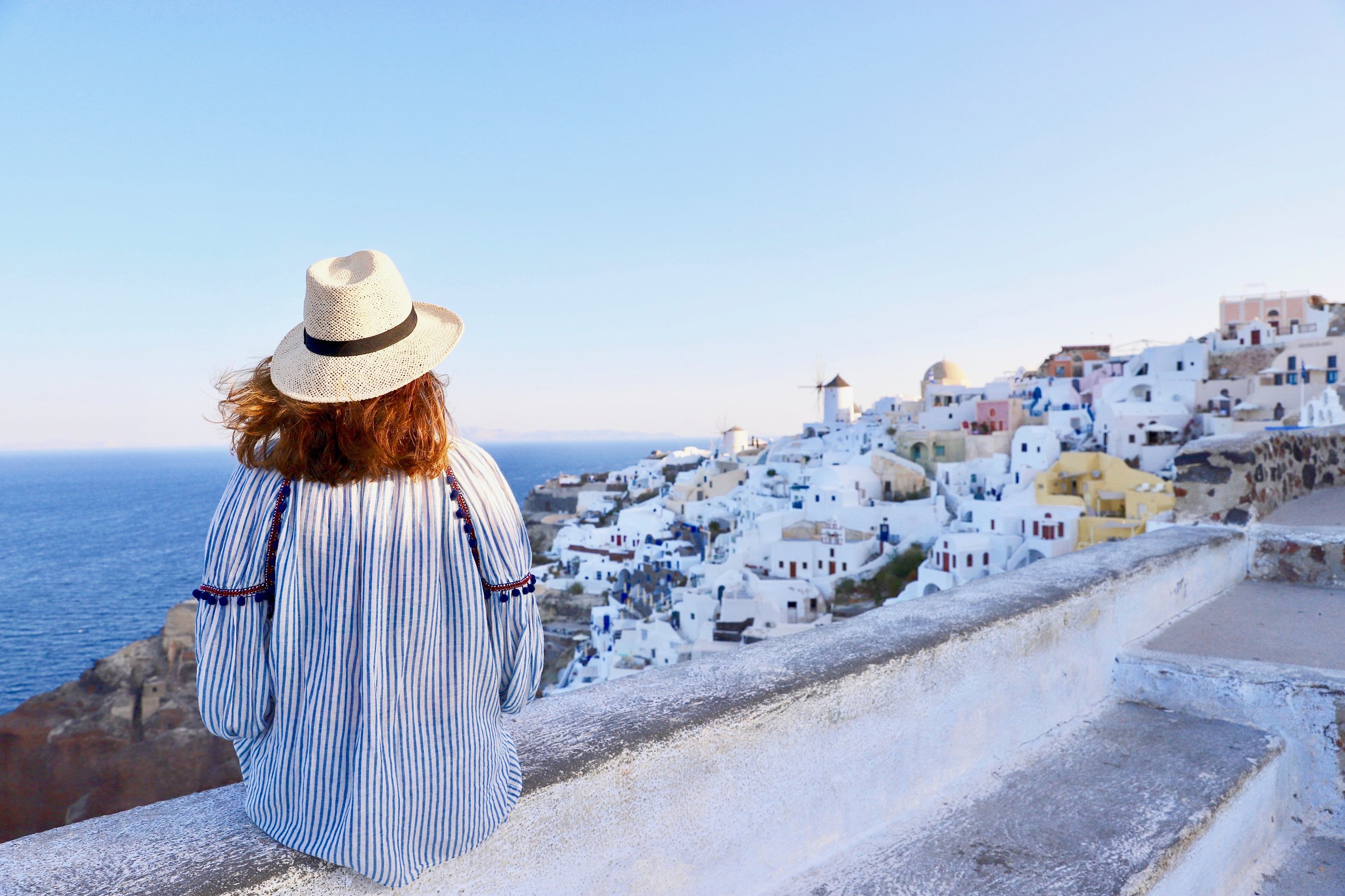 Dame med bluse i det greske flaggets farger og hatt som sitter på en mur og ser soloppgangen over Oia på øya Santorini. 