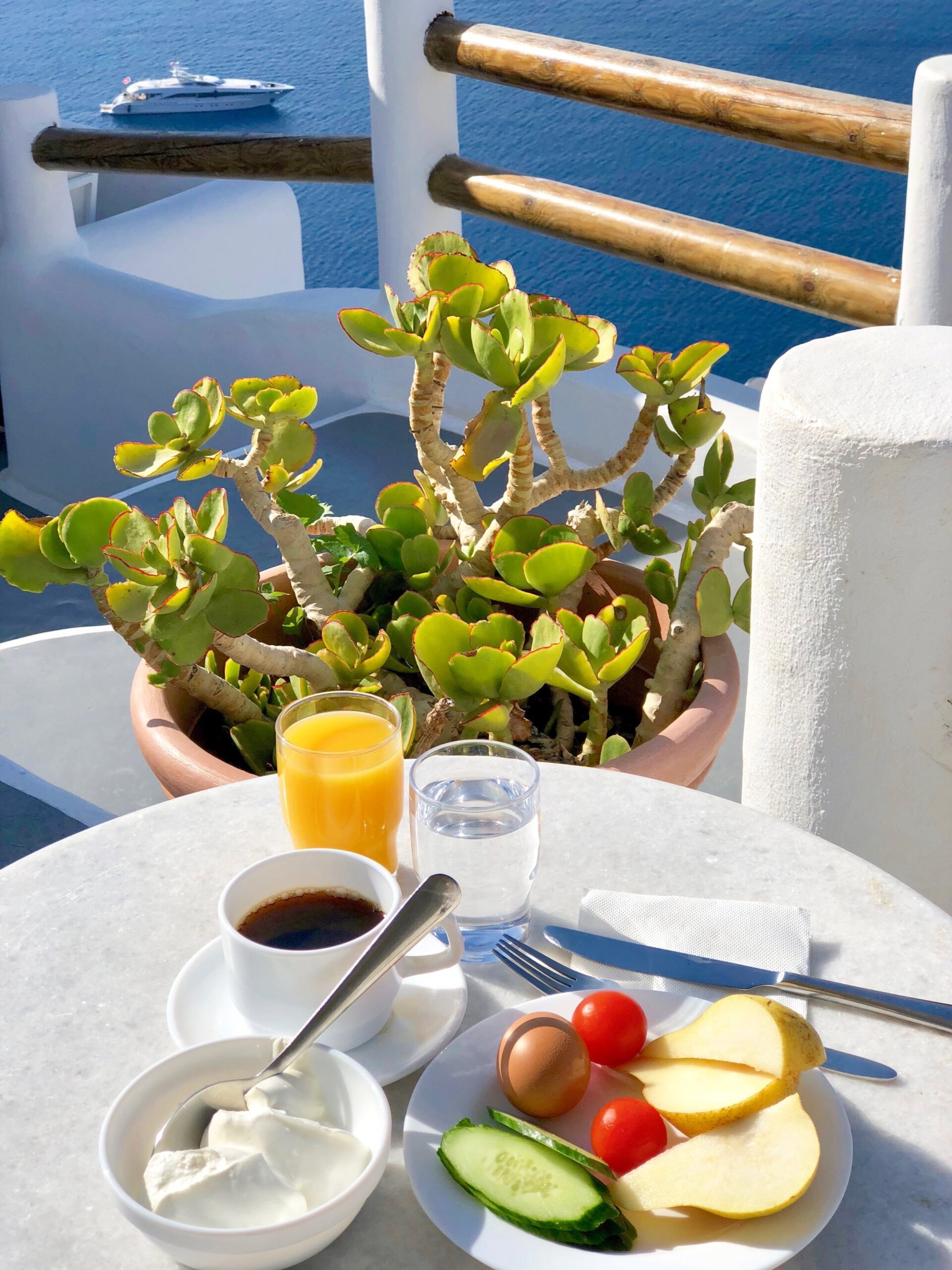 Frokost bestående av juice, kaffe, gresk yoghurt, egg, pære, tomat og agurk. I bakgrunnen en lekker cabincruiser på det blå havet. 