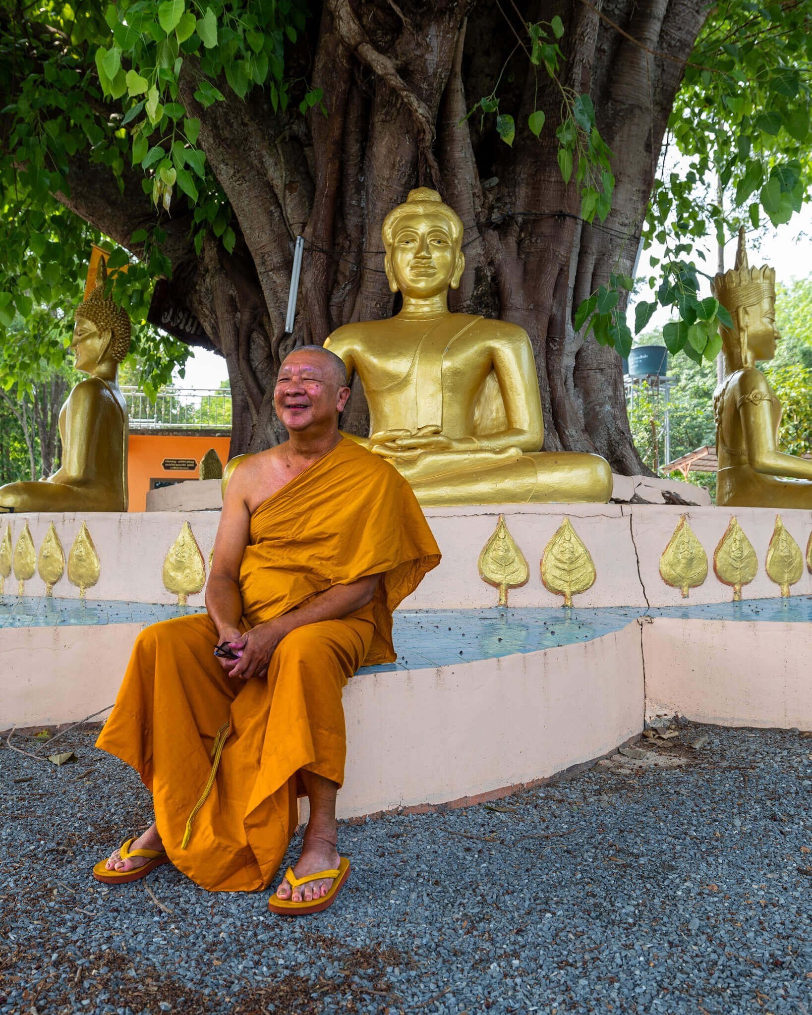 En smilende munk i orange drakt med orange sandaler foran en statue av buddha i gull.