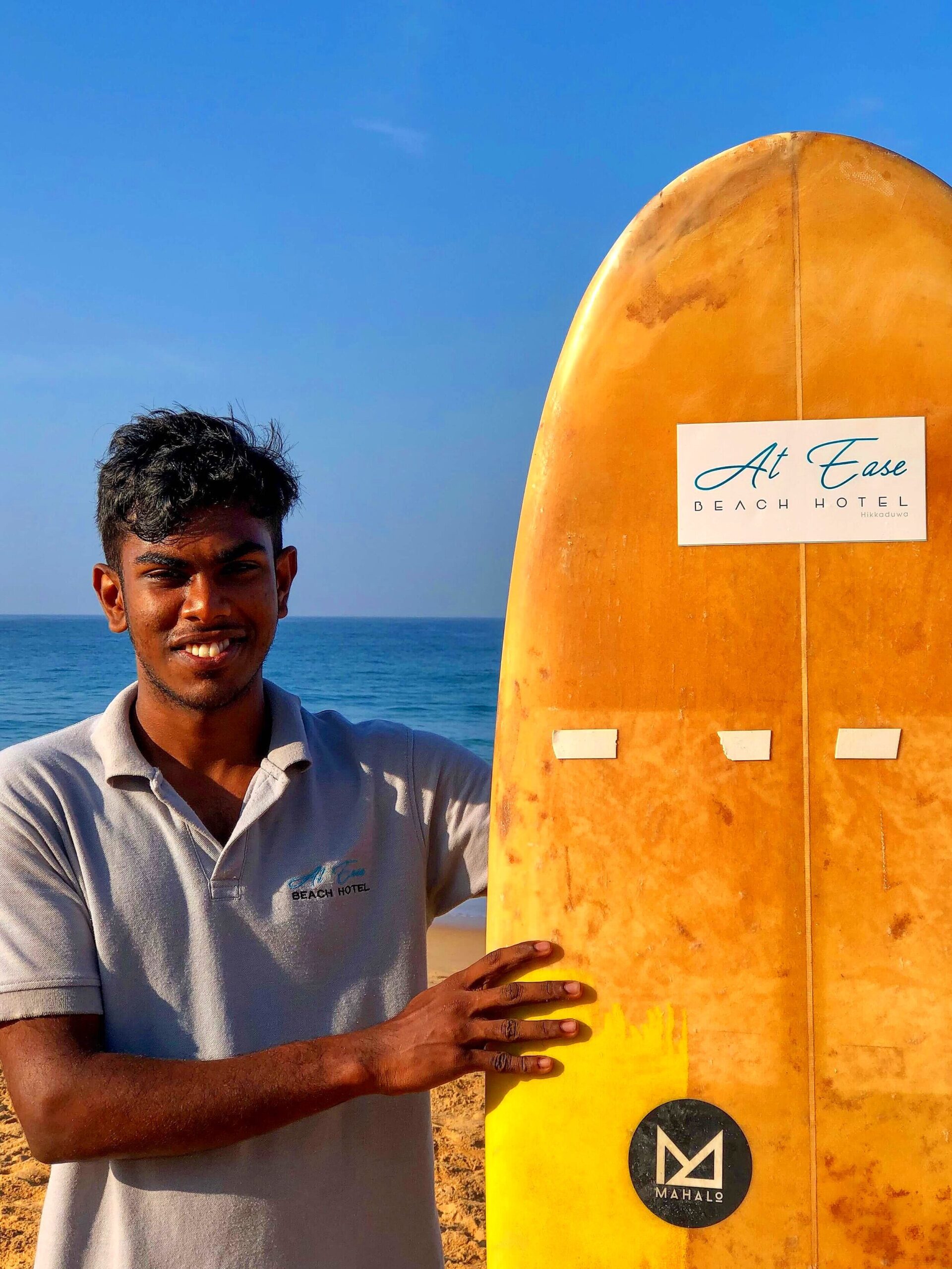 Smilende surfeinstruktør med surfebrett på stranden ved At Ease Beach Hotel Sri Lanka. Havet i bakgrunnen.
