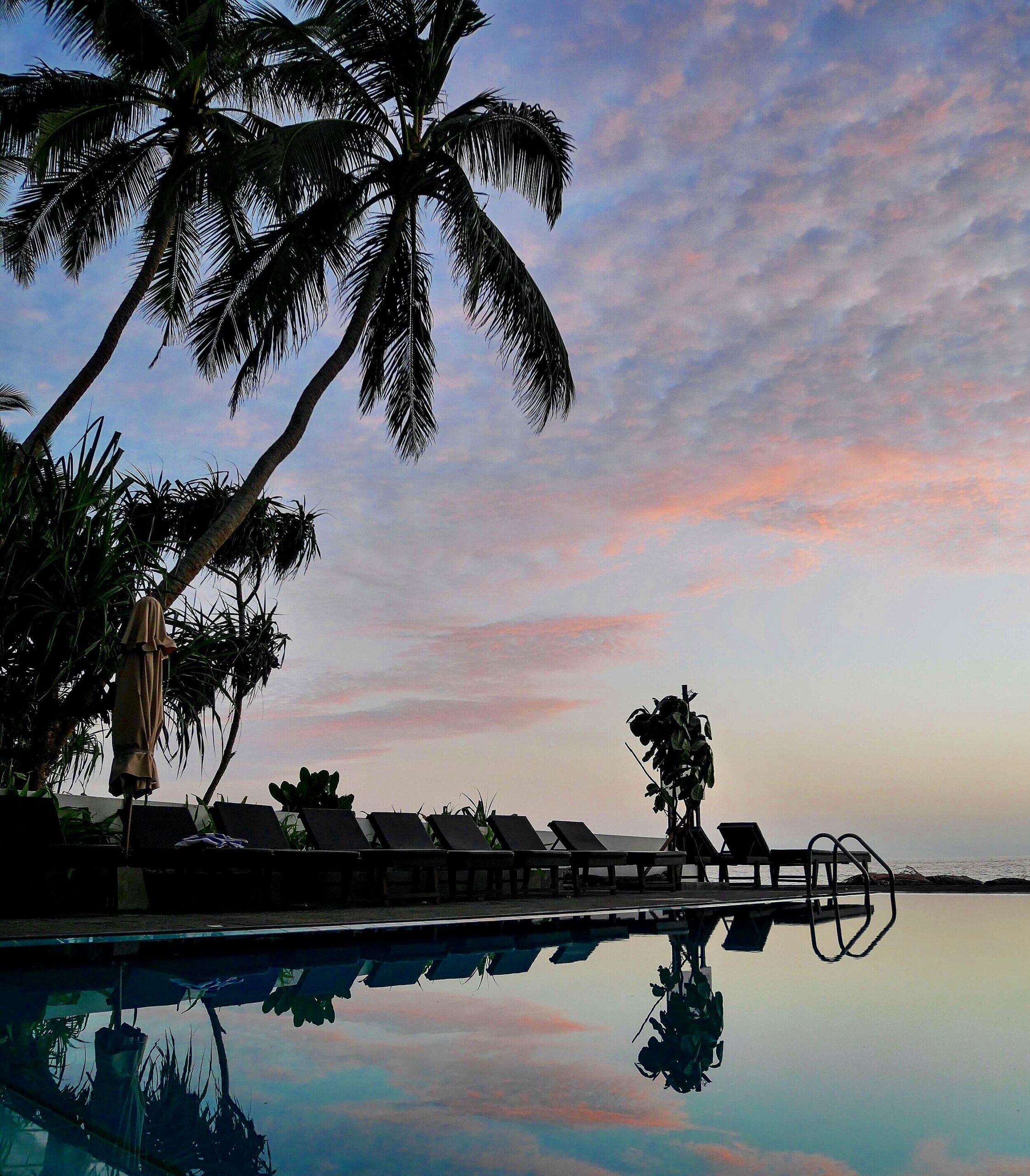 Solnedgang på At Ease Beach Hotel Hikkaduwa. På bildet ser du solstoler, parasoller og bassenget.