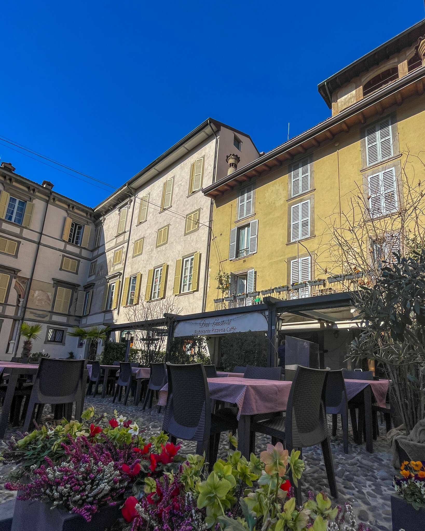 Sjarmerende restaurant med uteservering i Bergamo Italia. Her står bord og stoler ute mellom hyggelige gamle bygg med skodder foran vinduene. 