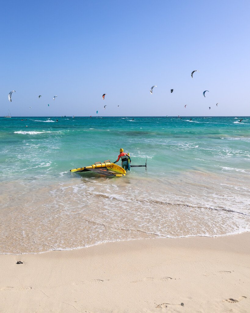 Strand full av kitere på Fuerteventura, Spanias surfe og kiteparadis. Dame på vei ut i sjøen med sin kite.