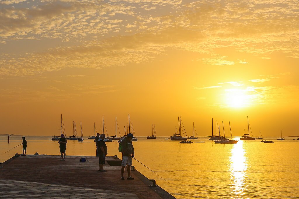 Soloppgang som farger himmelen gul i Corralejo på Kanariøyene. Solen speiler seg i vannet og over båtene. Folk står på bryggen og fisker.