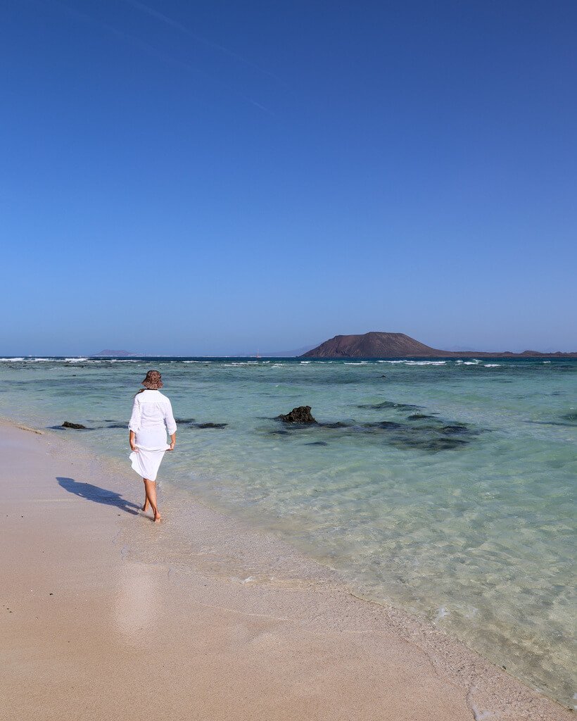 Strand på Fuerteventura i Spania. Dame i hvit kjole med hatt på hodet går barbeint bortover en hvit sandstrand. Havet er turkist og himmelen blå. 