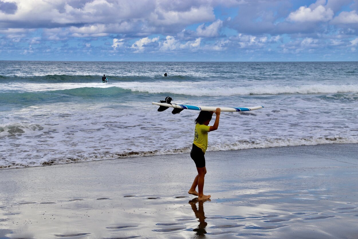 Jente med surfebrett på hodet på stranden Playa de Las Canteras i Las Palmas de Gran Canaria. Hav, himmel og surfere som venter på den perfekte bølgen.