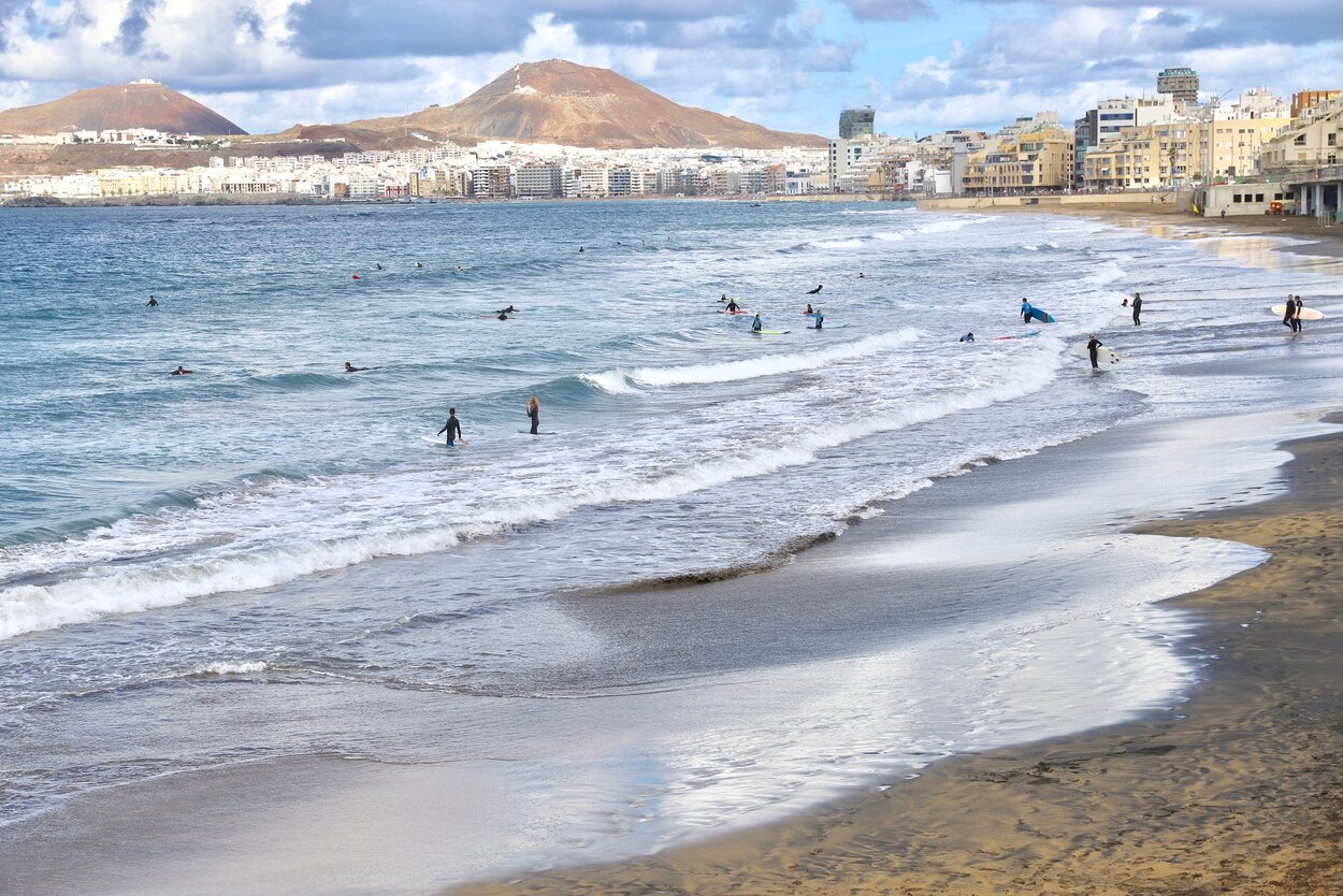 Utsikt over Las Canteras-stranden i Las Palmas, Gran Canaria, med surfere i vannet, byen i bakgrunnen og fjell som ruver i horisonten.