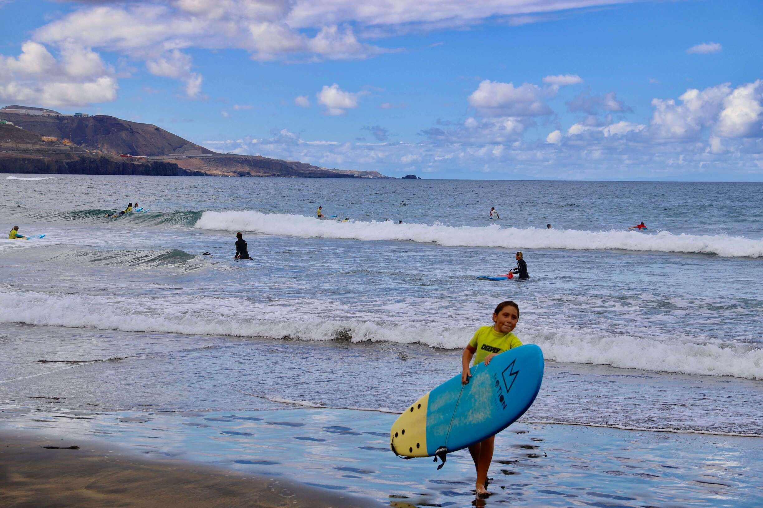 En jente med surfebrett på Playa de Las Canteras i Las Palmas, med surfere og det bla havet i bakgrunnen.