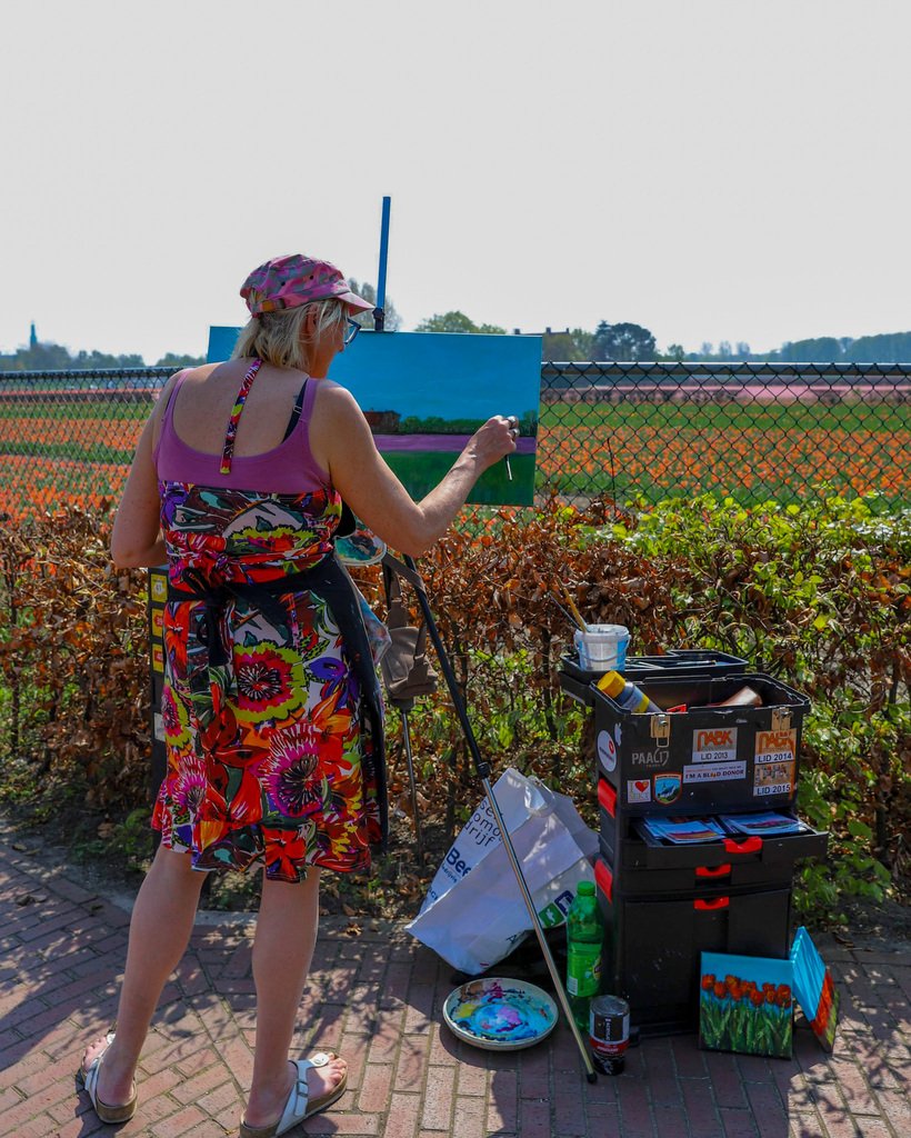 En kvinne star ved et staffeli i Lisse, Nederland, omgitt av et hav av blomstrende tulipaner i alle regnbuens farger. Hun har på seg en blomstrete sommerkjole og en caps som skjermer henne fra sollyset. Hendene hennes beveger seg fokusert over lerretet, hvor et detaljert bilde av tulipanene gradvis tar form.