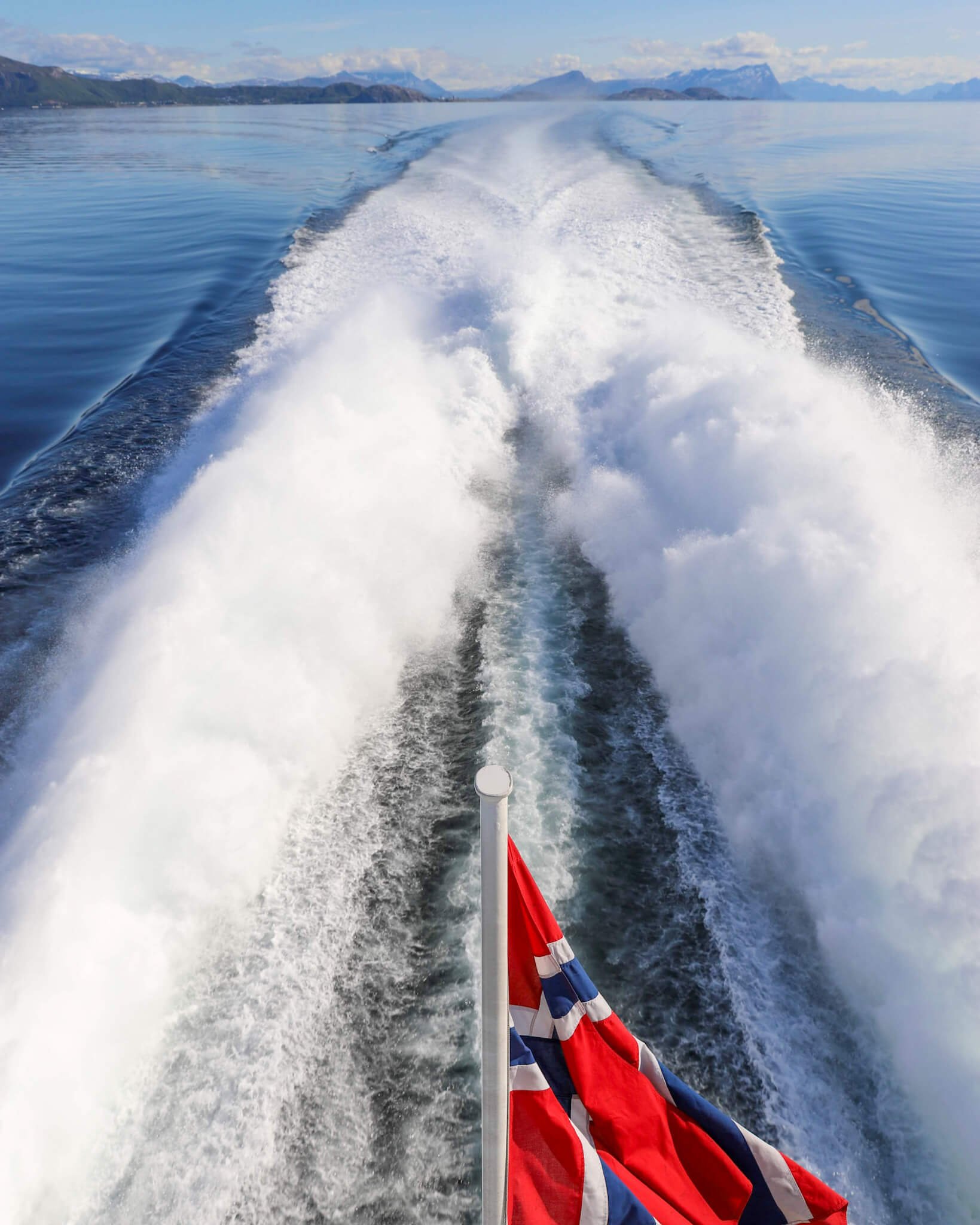 Bildet er tatt ombordstigning på hurtigbåt fra Helgelandskysten til Lofoten. Det norske flagget vaier i vinden når båten kjører og lager bølger på det blikk stille havet.