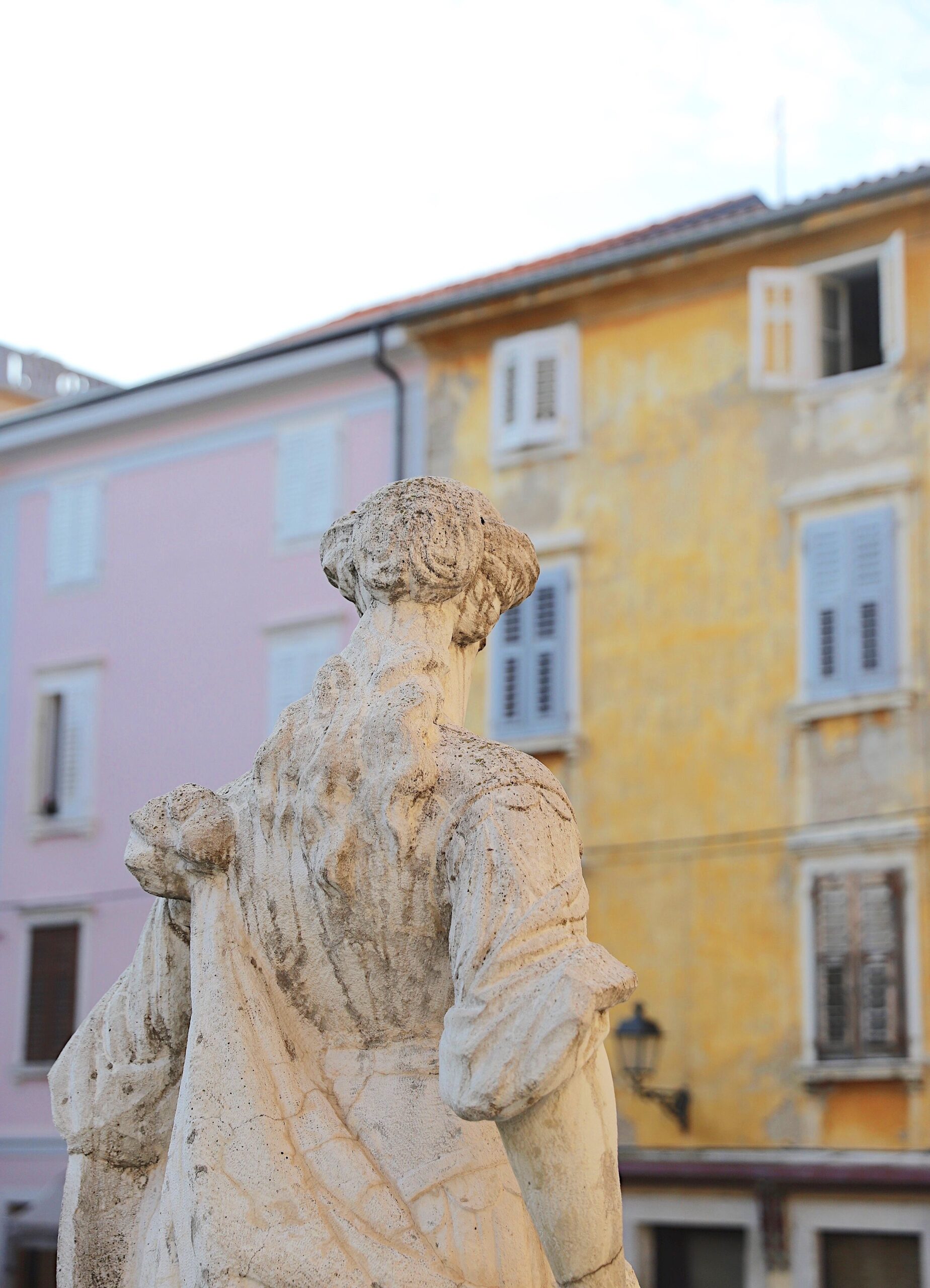 Statue av kvinne i Piran. I bakgrunnen et rosa og et gult bygg preget av tidens tann.