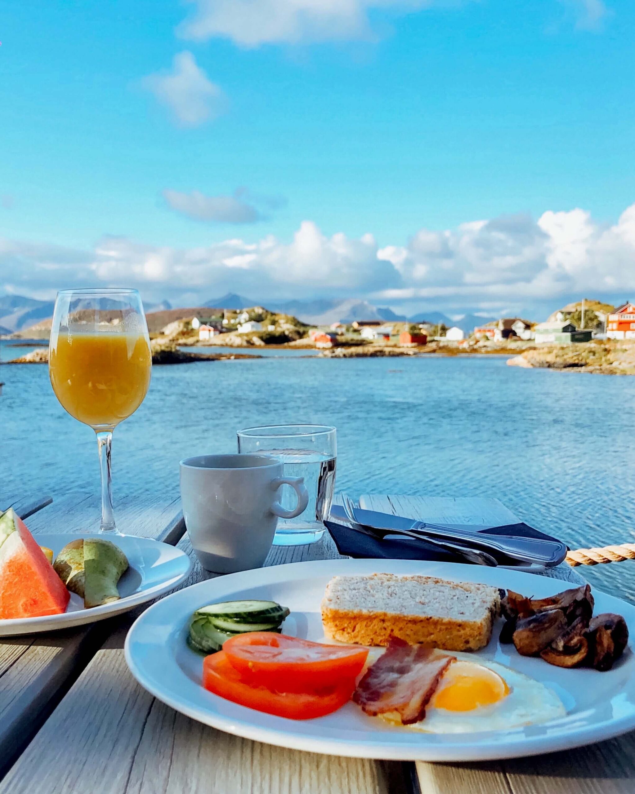 Frokost på Sommarøy Arctic Hotel. Kaffe, appelsinjuice, frukt, egg, bacon, brød, sopp, tomat og agurk. Frokosten inntas utenfor hotellet med utsikt over havet. 