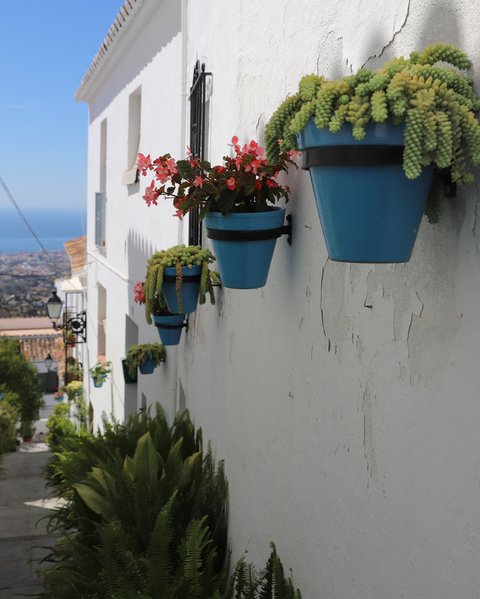 Mijas Pueblo i Spania. En hvit husvegg. På veggen henger det fargerike blomsterpotter med grønne planter og rosa blomster. Du ser sjøen herfra. 