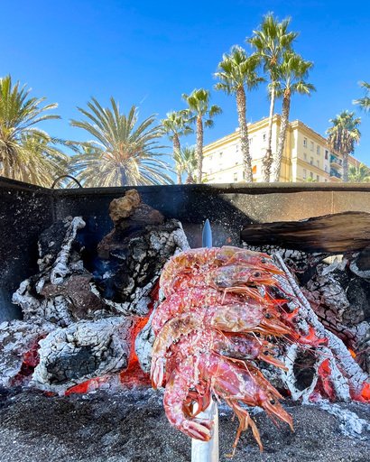 Reker grillet på spyd på kull på strand i Malaga. I bakgrunnen palmer og en vakker bygning. 