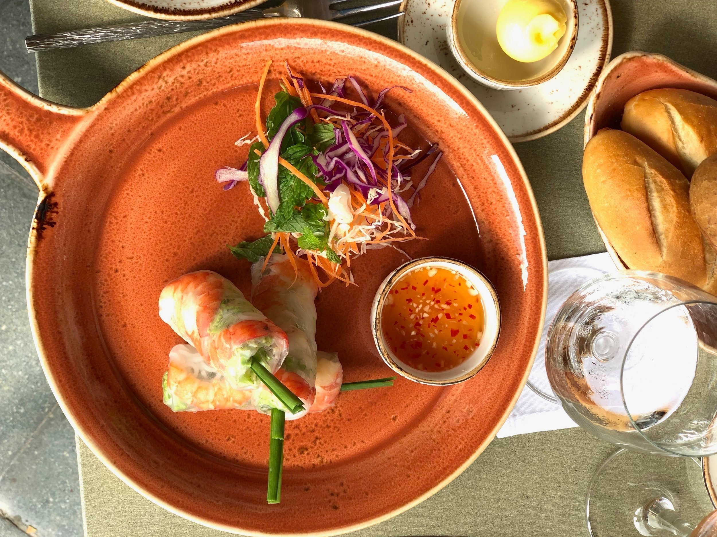 Vietnamese springrolls, salad, bread and water served on a rustic plate in colour orange in the restaurant. 