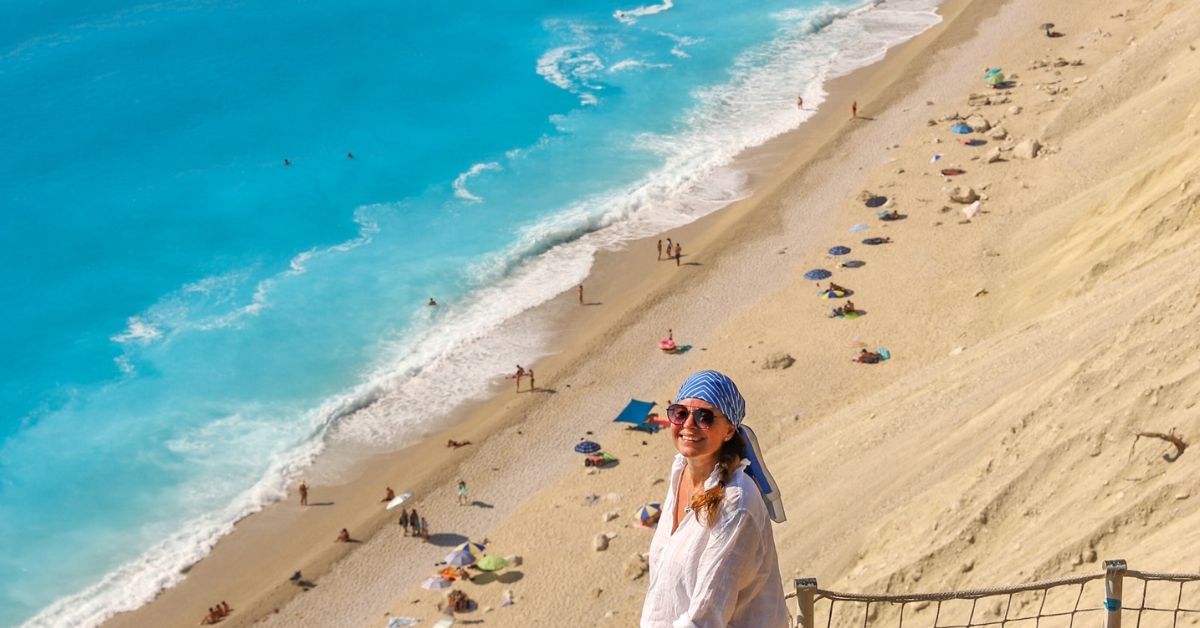 Merete Moen, World with a Suitcase, på strand på gresk øy med turkist hav.