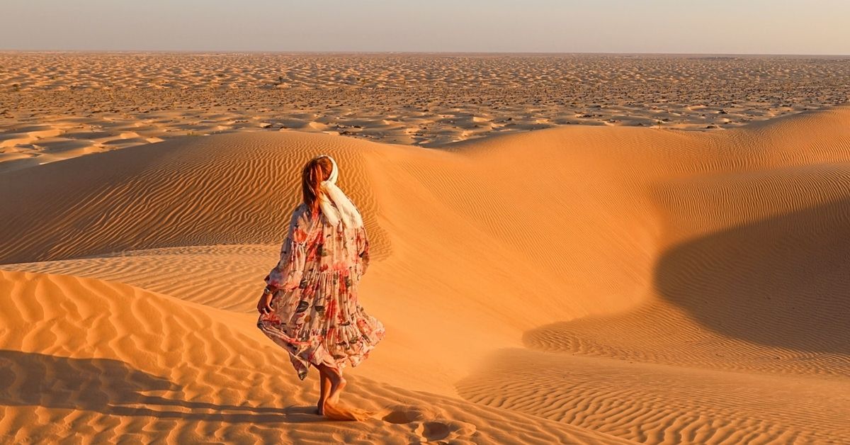 Dame med blomstrete kjole og turban på hodet, turist, vandrer barbent på sanddynene i Omans ørken ved solnedgang.