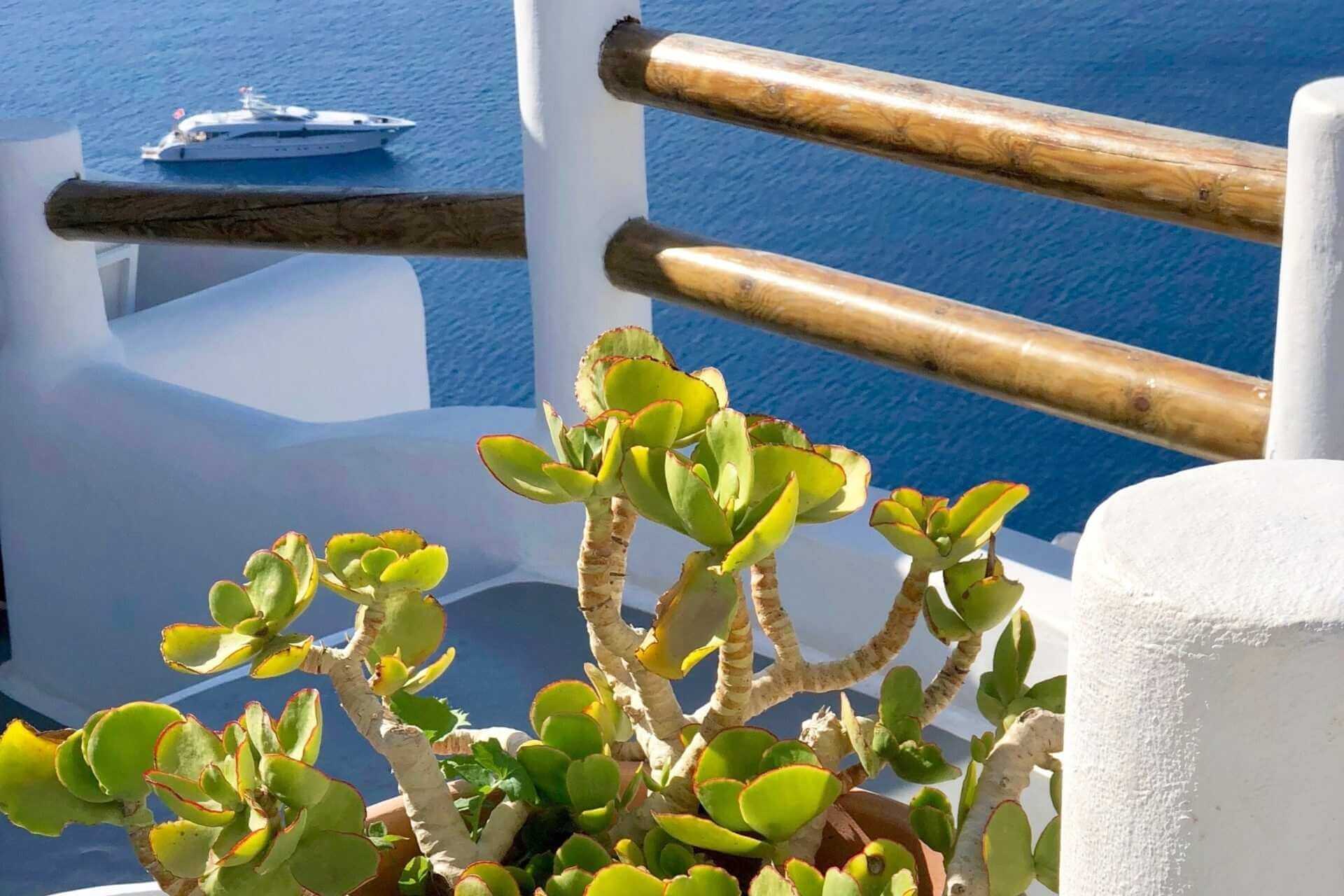 Utsikt over havet fra hvit balkong p&aring; Santorini, med gr&oslash;nn plante og yacht i bakgrunnen.