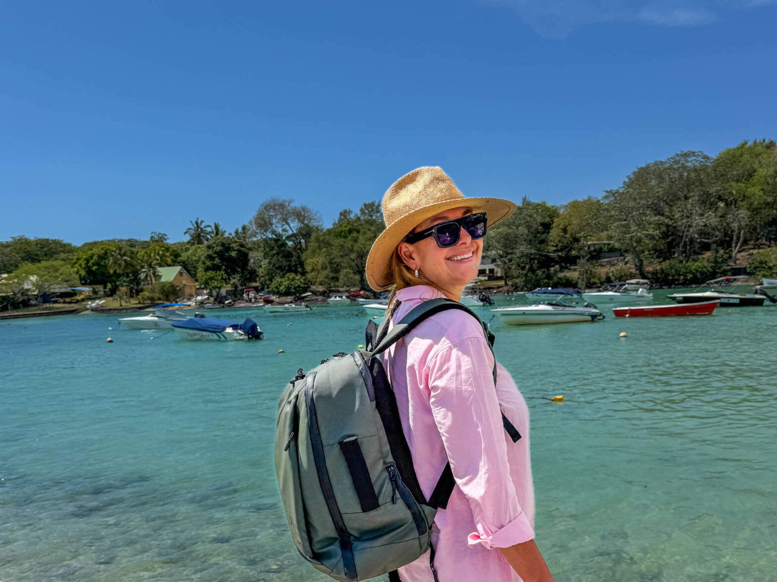 Smilende kvinne p&aring; reise ved sj&oslash;en med sekk p&aring; ryggen og hatt p&aring; hodet.