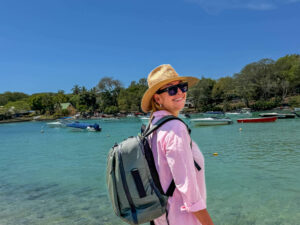 Smilende kvinne på reise ved sjøen med sekk på ryggen og hatt på hodet.