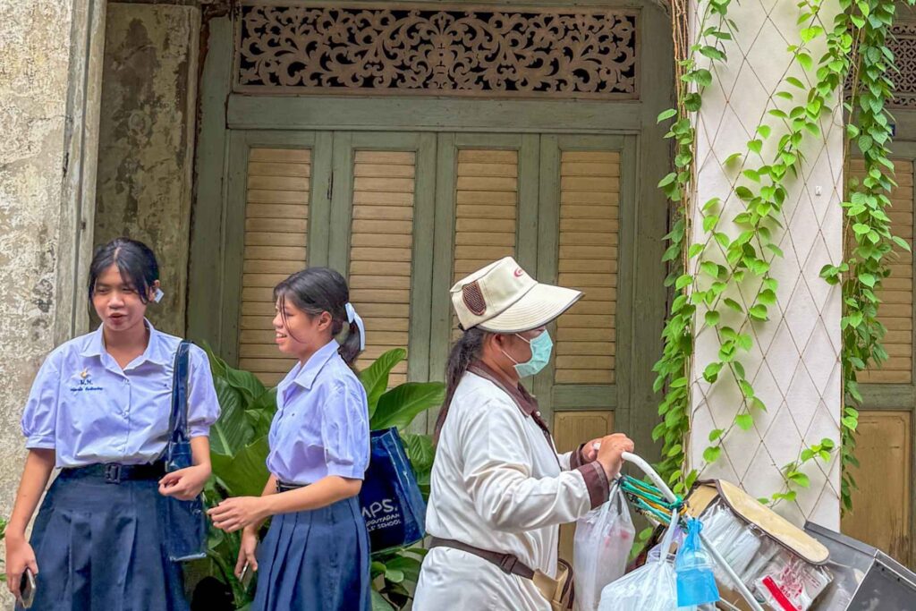 Hverdagsliv i Bangkok med skolejenter og gatefeier foran historisk bygning i gamlebyen.