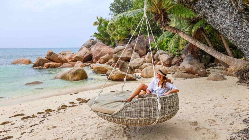 Kvinne på ferie med hatt og lyse klær slapper av i kurvstol som henger i en palme på Constance Lemuria på Seychellene.