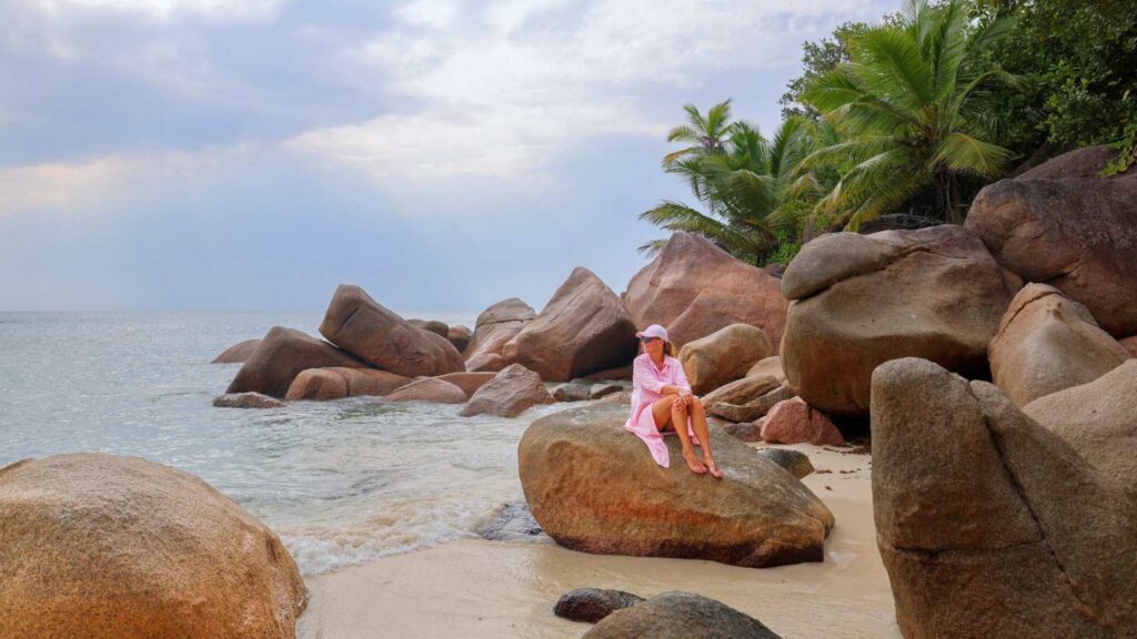 Same i rosa kjole sitter på granittstein på strand på Seychellene.