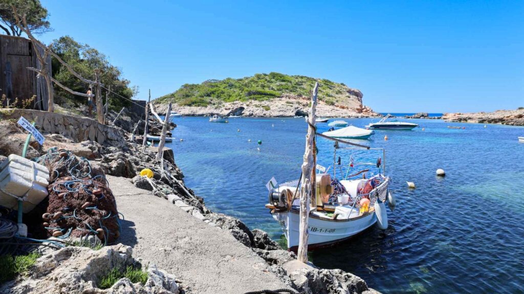 Tradisjonell fiskebåt og havn i Cala Portinatx nord på Ibiza med små båter, klart turkis hav og klipper langs kysten.