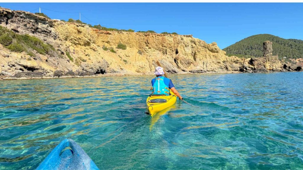 Kajakkpadling i krystallklart turkist vann ved Es Figueral i Santa Eulària på nordøstkysten av Ibiza omgitt av klipper og furuskog.