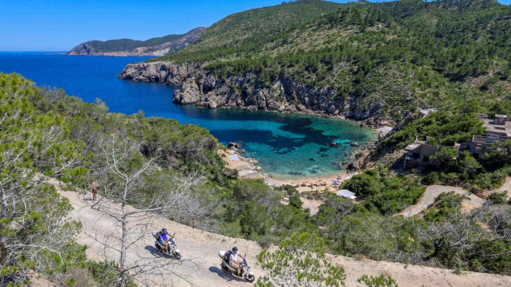 Utsikt over bukten i Sant Joan de Labritja på nordkysten av Ibiza med turkist hav, klipper, furutrær og den lille strandrestauranten Taverna nede ved vannet.