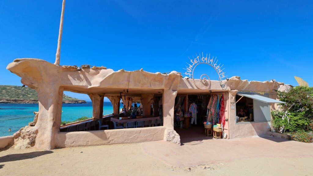 Sunset ved Cala Comte på Ibiza med utsikt over turkist hav, bohemsk arkitektur og restaurant helt ute ved klippene.