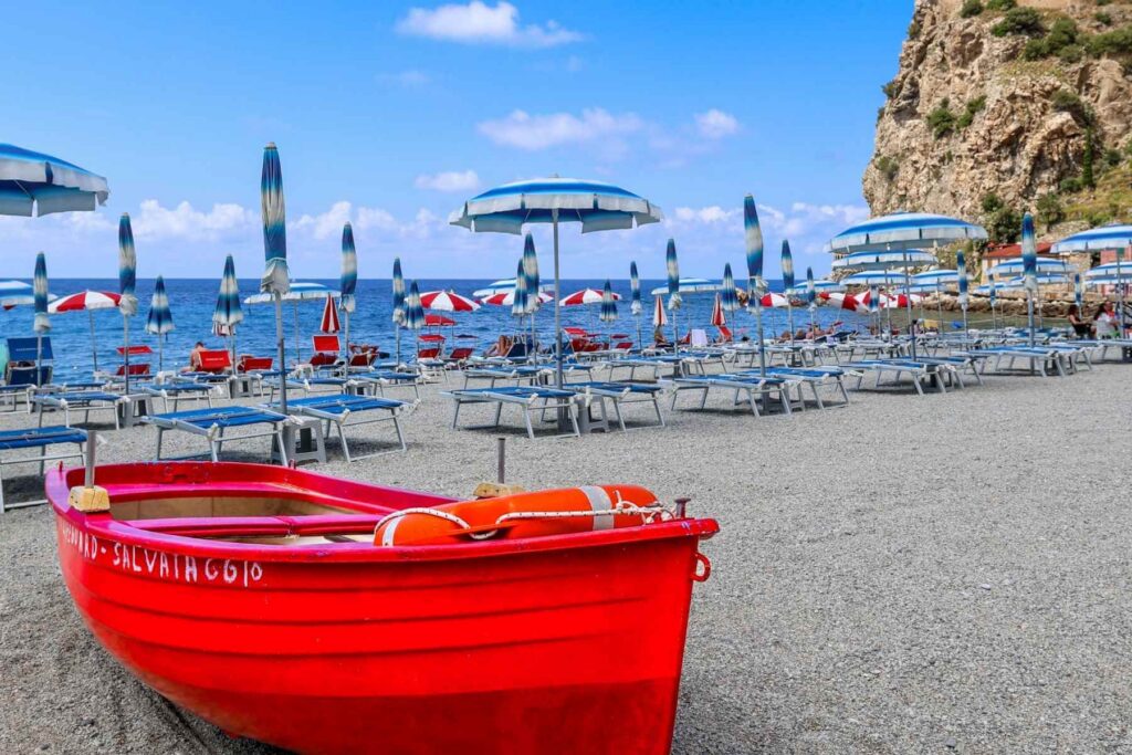 Rød robåt på steinstranden i Scilla, Calabria. I bakgrunnen solsenger og parasoller.