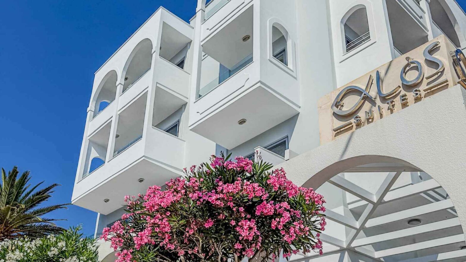 Alos Suites i Pigadia på Karpathos, hvit hotellbygning med balkonger og rosa blomster.