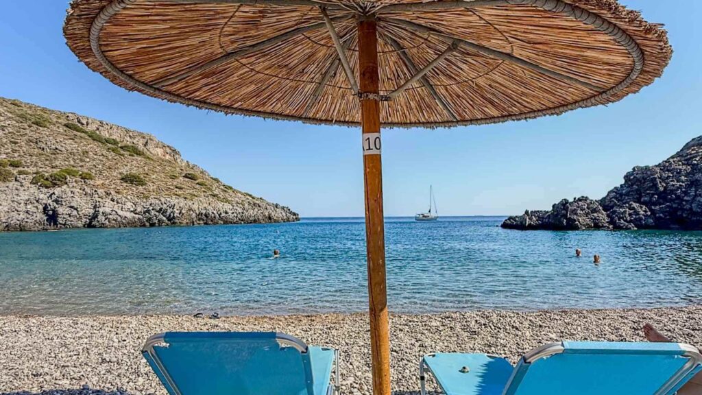Rullesteinstranden Chalkos Beach med str&aring;parasoll, bl&aring; solsenger og klart vann i en skjermet bukt.