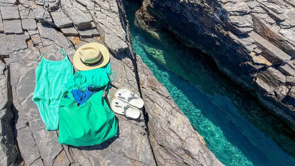 Turkis badedrakt, gr&oslash;nn strandveske, str&aring;hatt og sandaler p&aring; klippen ved krystallklart hav kjennetegner denne greske &oslash;ya.