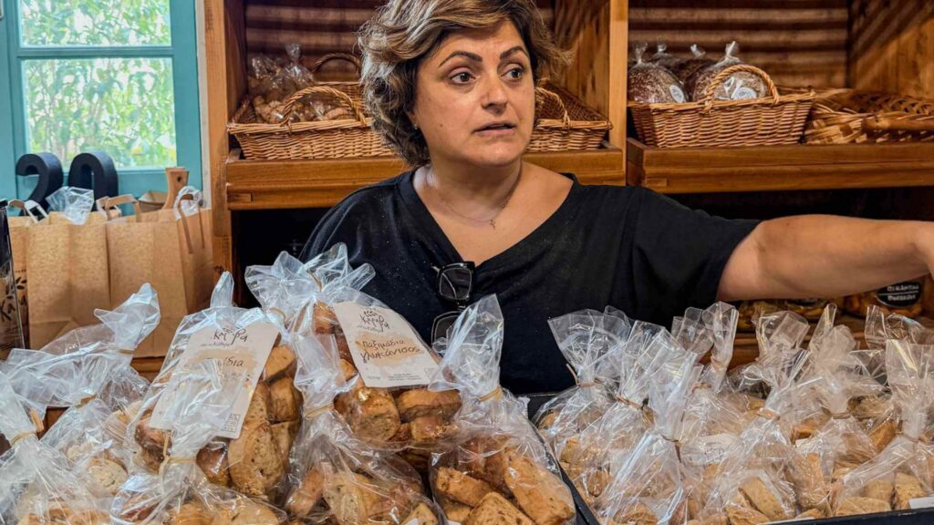 Kvinne i et tradisjonelt bakeri p&aring; omgitt av kurver og poser med lokale greske bakervarer.