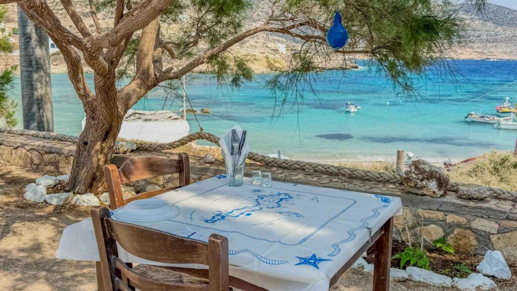 Tradisjonell gresk taverna ved sj&oslash;en i Lefkos p&aring; Karpathos, med trebord, skyggefulle tr&aelig;r og turkist badevann. 